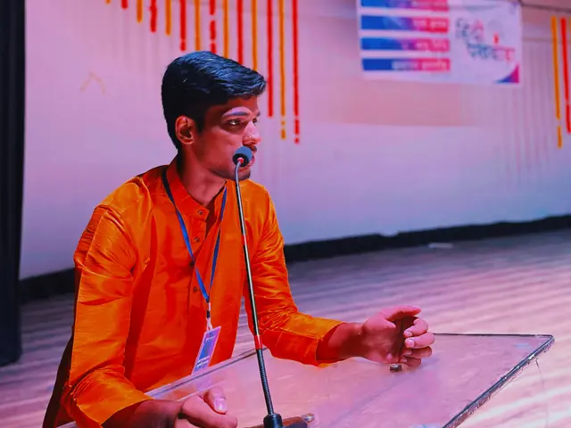 The image shows a man sitting at a podium with a microphone in front of him, wearing an orange...