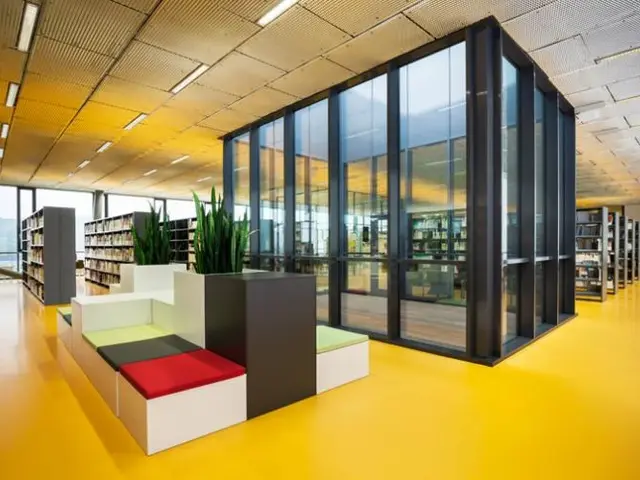 The image shows a large room filled with lots of bookshelves, a sofa set, houseplants, glass doors,...