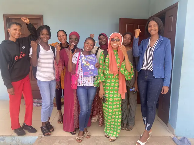 The image shows a group of women standing next to each other in front of a building, smiling and...