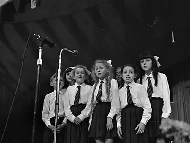 The image shows a group of young girls standing on top of a stage, singing into microphones with...