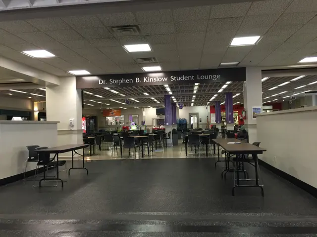 The image shows a large room filled with lots of tables and chairs, pillars, a name board, lights...