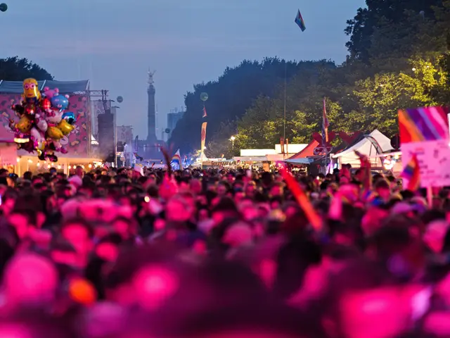The image shows a large crowd of people at a music festival at night, illuminated by colorful...