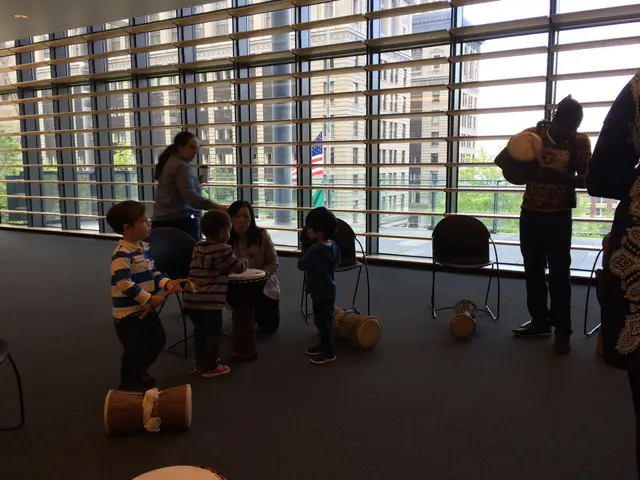 The image shows a group of children playing drums in front of a large window, with chairs scattered...