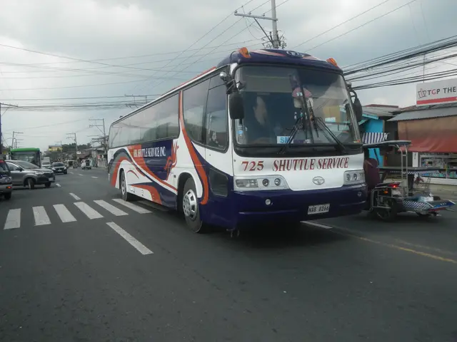 The image shows a shuttle bus driving down a street lined with tall buildings, electric poles with...